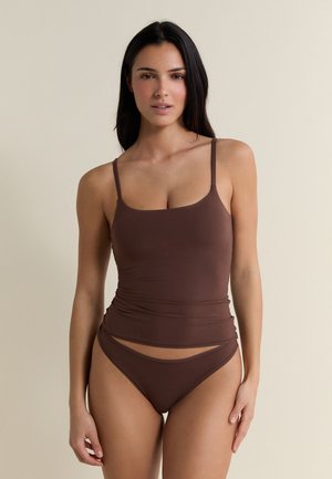 Donna con lunghi capelli scuri che indossa un top camisole marrone e slip bikini abbinati, in piedi davanti a uno sfondo beige.