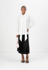 White long-sleeve blouse with a tied collar, paired with a black pleated skirt. Accessories include a small pink handbag and black strappy heels.