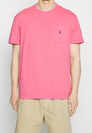 Mężczyzna ubrany w jaskraworóżową koszulkę z krótkim rękawem z małym granatowym logo na kieszeni na piersi oraz beżowe szorty.