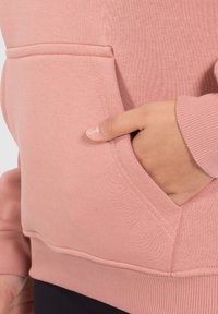 Sweatshirt en coton rose avec une poche kangourou, des poignets côtelés et une texture lisse, présenté avec une main reposant à l'intérieur de la poche.