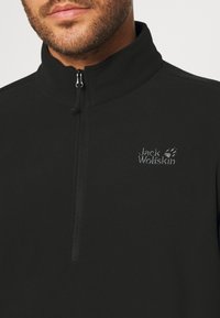 Schwarzer Fleece-Pullover mit hohem Kragen und Teilreißverschluss. Auf der linken Brust ist das "Jack Wolfskin"-Logo in Grau gestickt.