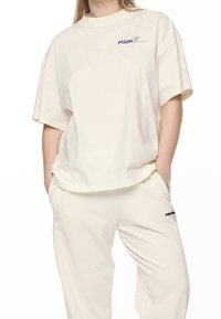 Krémové oversized tričko z měkké bavlny, s krátkými rukávy a modrým logem PGDR; v kombinaci s odpovídajícími krémovými tepláky.