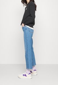 Sudadera negra, jeans de tiro alto de un azul claro, zapatillas blancas y moradas. Con mangas enrolladas y detalle del logo en la sudadera. Textura plana.
