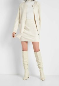Veste en similicuir crème à coupe droite, mini-jupe assortie et bottes montantes jusqu'aux genoux ; texture lisse avec des accents métalliques minimalistes.