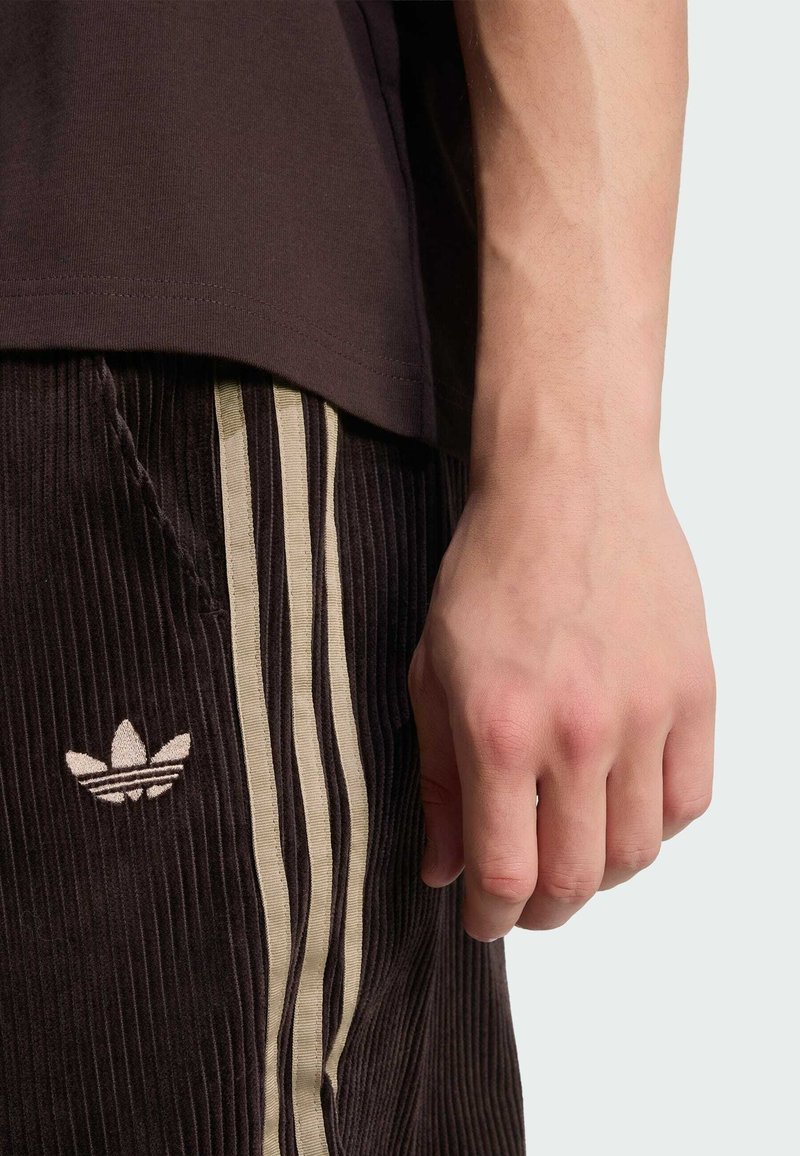 Højre hånd hviler ved siden af mørkebrune kordfløjlsbukser med beige Adidas-logo og tre striber, iført en sort skjorte.