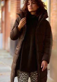 Manteau rembourré marron avec capuche, fermeture éclair à l'avant, et un pull noir à côtes en dessous. Associé à des leggings à motifs avec un imprimé léopard.