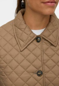 Veste beige matelassée avec un grand col, présentant des motifs en diamant et une fermeture à boutons. Texture lisse, avec des détails de boutons et de couture noirs.