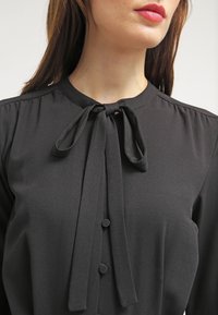 Blouse noire avec un col à nouer, dotée d'une texture lisse, d'un col rond et de deux boutons à l'avant.
