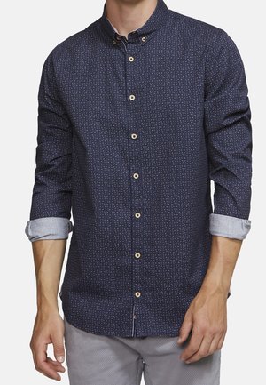 Hombre con camisa azul marino abotonada con pequeños puntos multicolores, mangas remangadas, combinado con pantalones gris claro, de pie frente a un fondo blanco.