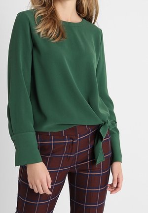 Vrouw draagt een groene blouse met lange mouwen en een zijstrik, en een bruine geruite broek met blauwe en rode strepen.