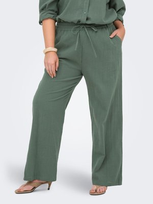 Femme portant un pantalon vert ample avec une taille à cordon et la main dans la poche, associée à des sandales à talons beiges et un haut vert assorti.