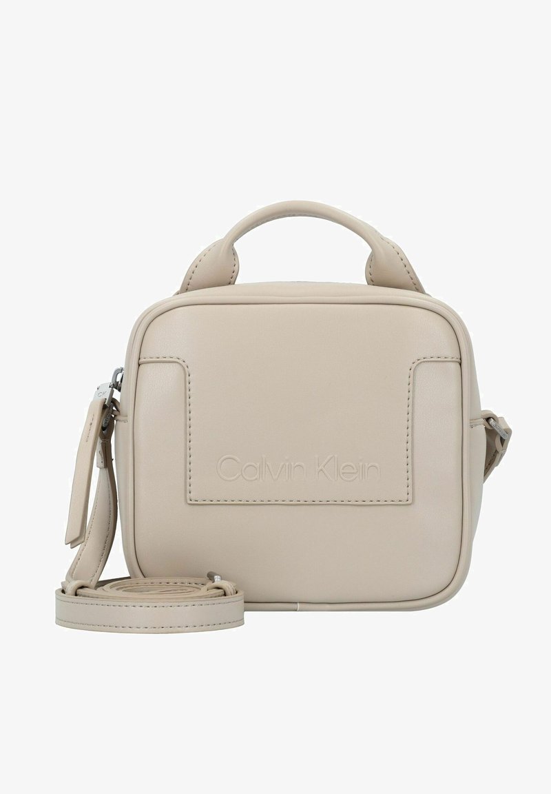 Petit sac bandoulière beige Calvin Klein avec poignée supérieure, poche avant et sangle d'épaule ajustable enroulée à la base.