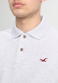 Světle šedé polo triko s jemnou texturou, kontrastním červeným vyšitým logem a třemi hnědými knoflíky na légu.