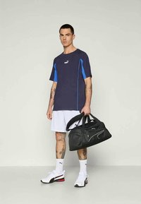 Tumesinine spordipoolkurtiga sinise aktsendiga, mis on ühendatud valgete lühikesed pükstega. Must sportkot ja valged tennisetossud punaste aktsentidega. Näha on tätoveeringud.