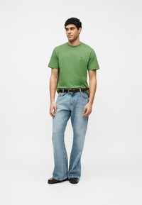 Άνδρας που φοράει πράσινο κοντομάνικο πουκάμισο, ανοιχτό μπλε τζιν παντελόνι με φαρδιά μπατζάκια και καφέ πλεκτή ζώνη, με σκούρα καφέ παπούτσια, στέκεται μπροστά σε λευκό φόντο.