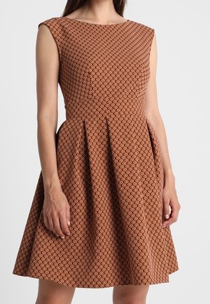 Mouwloze A-lijn jurk met bruin- en oranje diamantpatroon, getailleerde taille en boothals, gedragen door een staande vrouw.