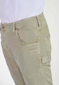 Lichtbeige cargobroek met meerdere zakken, gecombineerd met een wit ingestopt shirt, gezien vanaf de taille naar beneden vanuit een zijaanzicht.