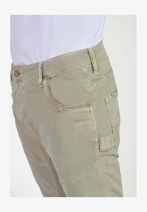 Pantalon cargo beige clair avec plusieurs poches, porté avec une chemise blanche rentrée, vu de la taille vers le bas à un angle latéral.
