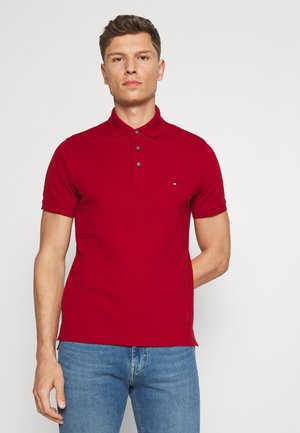 Άντρας που φοράει κόκκινο κοντομάνικο πόλο με κουμπιά και ανοιχτόχρωμο μπλε τζιν, στέκεται με ουδέτερη έκφραση.