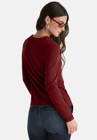 Frau mit langen dunklen Haaren trägt Schildkrötensonnenbrille, silberne Creolen, ein burgunderrotes Langarmoberteil und dunkelblaue Jeans und blickt zur Seite.