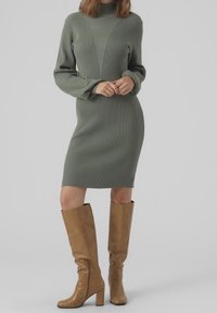 Robe côtelée mi-longue de couleur verte douce avec un col montant et des coutures verticales ; associée à des bottes en cuir marron clair montant jusqu'aux genoux.