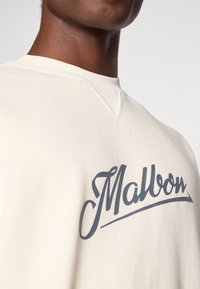 Krémová mikina s okrúhlym výstrihom, s výrazným tmavošedým logom "Malbon" cez hrudník a viditeľnými švovými detailmi.