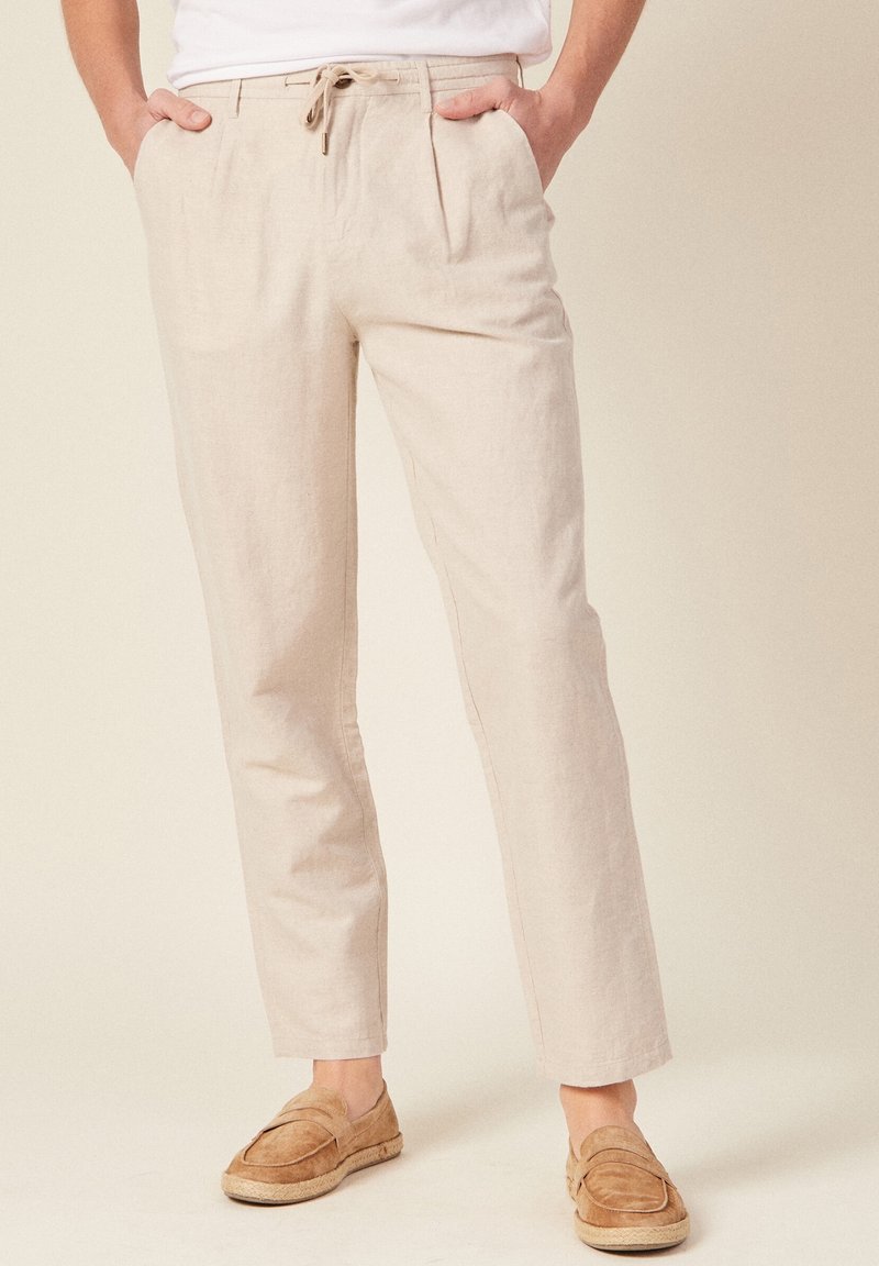 Light beige linen trousers with a drawstring waist and pleated front. Styled with flat, beige loafers for a relaxed look.