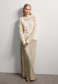 Suéter de punto crema con patrón de diamantes y mangas largas; falda maxi beige con bordado de hojas de ginkgo; combinada con sandalias en tono marrón.