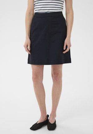 Žena oblečená v námorníckej modrej sukni A-rovného strihu, pruhovanom tielku bez rukávov a čiernych sieťovaných nazúvacích topánkach, stojaca na jednoduchom bielom pozadí.