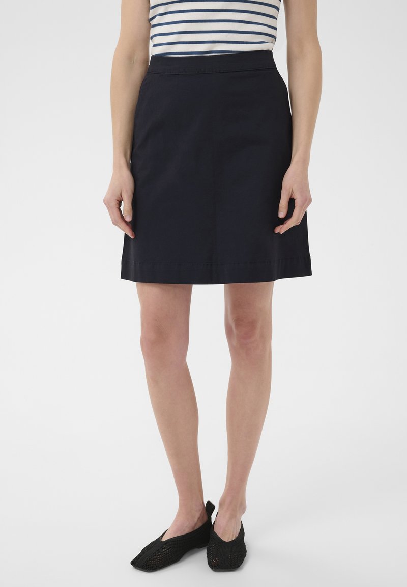 Femme portant une jupe évasée bleu marine, un haut sans manches rayé et des chaussures à enfiler en mesh noir, debout devant un fond blanc uni.
