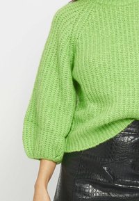 Helle grüne, gestrickte Pullover mit hohem Kragen und puffärmeln. Der strukturierte Stoff kontrastiert mit einem glänzenden schwarzen Rock im Krokodil-Design.