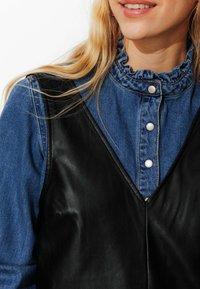 Gilet en cuir noir sur une chemise en denim bleu avec un col volanté et des boutons en argent, présentant une texture lisse et un design en V.