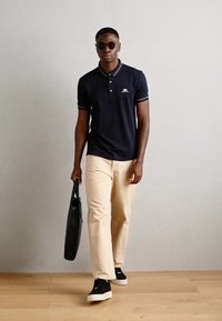 Marineblauwe poloshirt met witte accenten, beige chinos en zwarte sneakers. Zwarte tas in één hand. Gladde textuur, casual pasvorm.