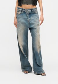 Γυναίκα που φοράει φαρδιά, ξεβαμμένα, φθαρμένα μπλε τζιν με ξεφτισμένα τελειώματα και πέδιλα ανοιχτής μύτης, μαύρο κοντό τοπ και μαύρο κοντό μπουφάν.