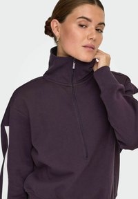Fioletowa bluza rozpinana z wysokim kołnierzem, o luźnym kroju, z kontrastowymi białymi wstawkami po bokach, wykonana z miękkiego materiału.