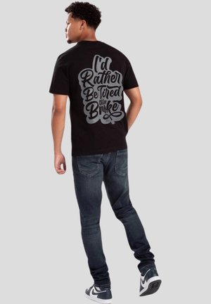 Camiseta negra con un gran gráfico gris en la parte de atrás que dice "Preferiría estar cansado que en quiebra". Combinada con jeans oscuros y zapatillas.