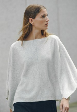 Ung kvinde med langt hår iført en løs lysegrå sweater og mørke bukser, der kigger til venstre mod en ensartet baggrund.