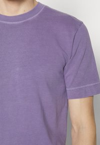 Fioletowa bawełniana koszulka z okrągłym dekoltem, krótkimi rękawami i subtelną teksturą. Posiada kontrastowe przeszycia wokół dekoltu i rękawów.