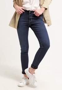 Jeans ajustados en denim azul oscuro con una textura suave, con una cintura de altura media y dobladillos acampanados. Combinados con zapatillas blancas.