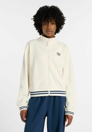 Personne aux cheveux bouclés portant une veste blanche zippée avec des poignets et un ourlet rayés bleus, associée à un pantalon bleu ample.