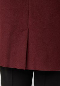 Bordeaux wollen jas met een gestructureerde afwerking, voorzien van een rechte zoom en een zijsplitdetail, gecombineerd met zwarte broek.