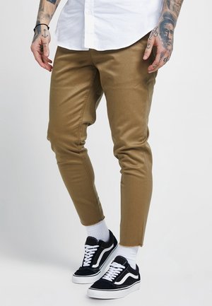 Pantaloni tapered color beige realizzati in misto cotone, caratterizzati da una finitura liscia. Abbinati a sneaker nere Vans e calze bianche alla caviglia.