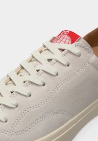 Ανοιχτό μπεζ αθλητικό παπούτσι από σουέτ με λευκά κορδόνια και κόκκινη ετικέτα στη γλώσσα με λευκό λογότυπο κόσμου και ματιών.