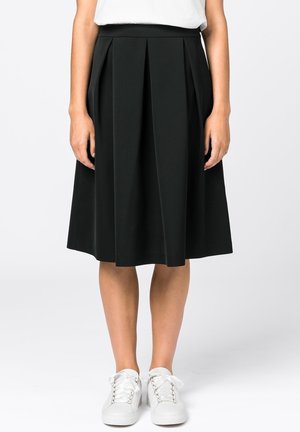 Femme portant une jupe plissée noire mi-longue associée à des baskets blanches, debout devant un fond clair uni.