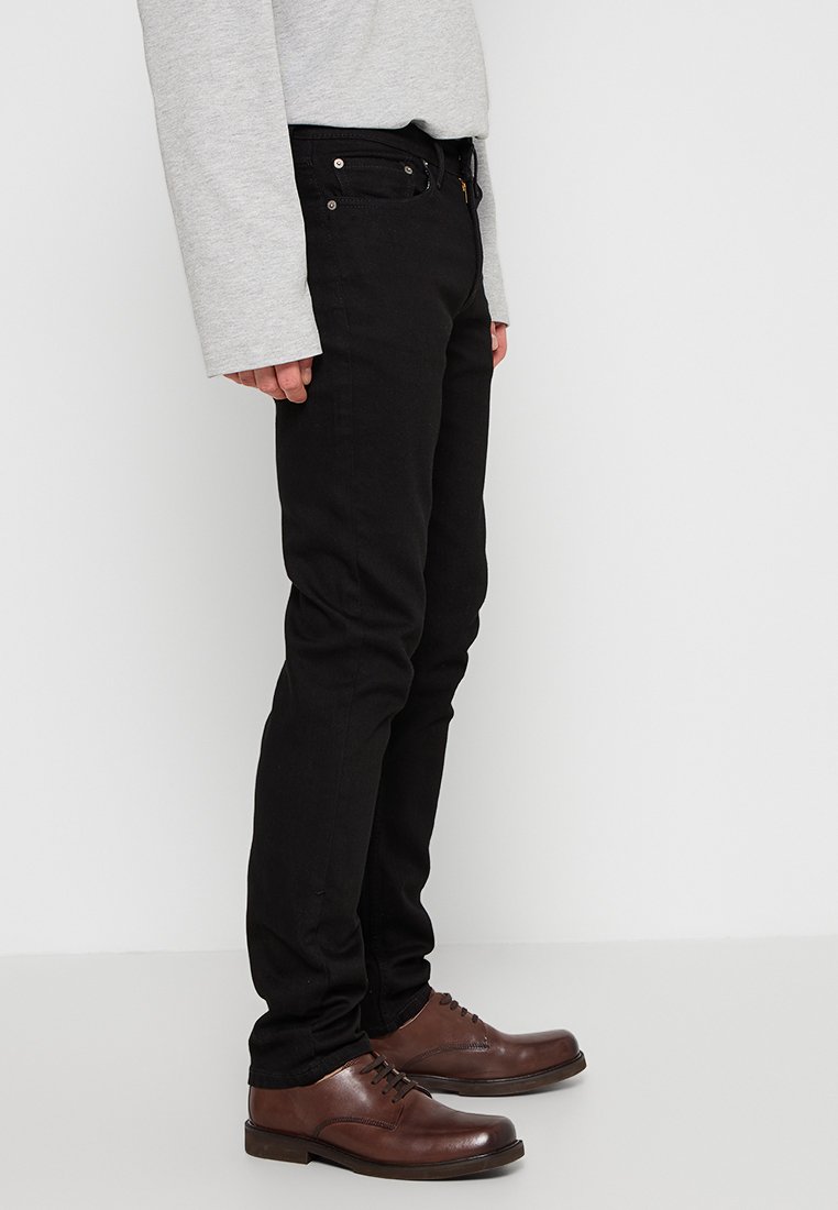 Persona que lleva pantalones ajustados negros, zapatos de cuero marrones con cordones y una camiseta de manga larga gris claro, de pie de lado sobre un fondo liso.
