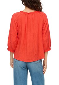 Blouse rouge texturée avec des manches 3/4 et un col froncé, associée à un jean bleu ample. Le tissu semble léger.
