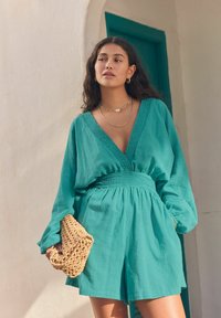 Robe rouge en teinte sarcelle à manches longues, décolleté en V profond, taille smockée et coupe décontractée. Tient une pochette beige texturée. Colliers en or superposés simples.