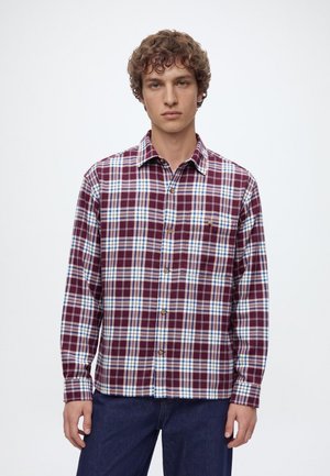 Un homme porte une chemise boutonnée à carreaux bordeaux, blancs et bleus avec un jean foncé, sur un fond clair uni.