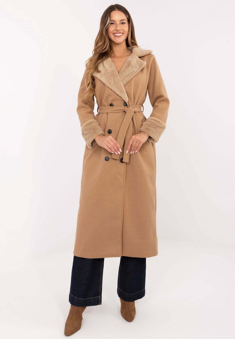 Langer hellbrauner Mantel mit Pelzkragen und Pelzmanschetten; verfügt über einen Wickelstil und eine doppelte Knopfreihe. Kombiniert mit ausgestellten dunklen Jeans.