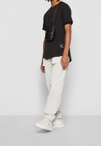 Osoba stojąca w czarnej koszulce, jasnobeżowych spodniach cargo, białych sneakerach i czarnej torbie na ramię na tle jednolitego, jasnego tła.